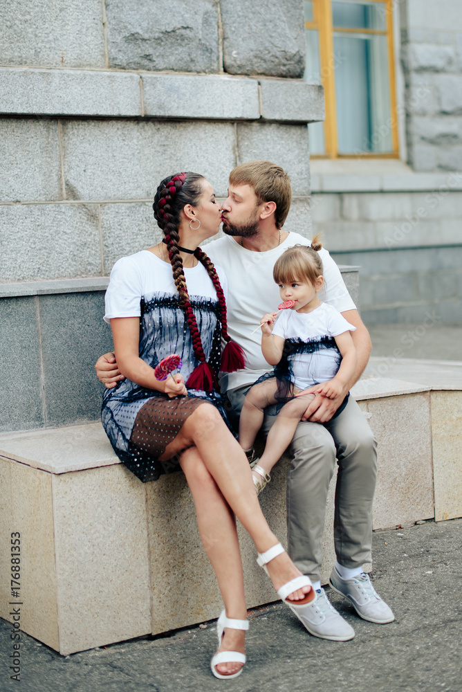 Young happy family with a little beautiful baby with blue eyes, with the grey of the building. Mother and daughter in matching dresses with candy
