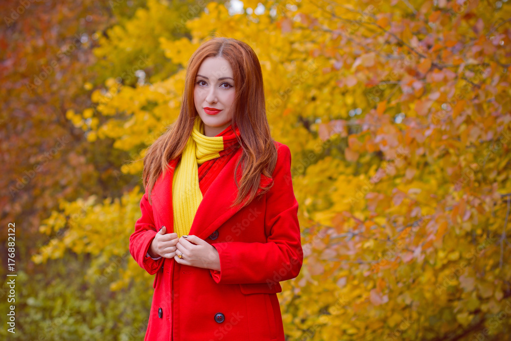 Cute and nice young beautiful woman in a red casual coat and red-yellow scarf walks in autumn city park. Pretty view of walking woman outside Cute and nice young beautiful woman in a red casual coat and red-yellow scarf walks in autumn city park. Pretty view of walking woman outside
