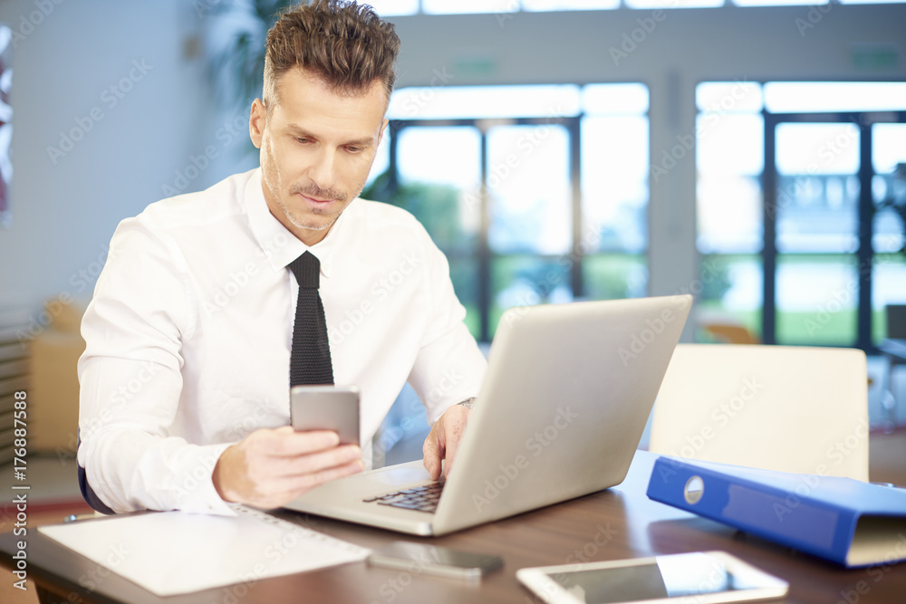 Businessman working at the office. Shot of a middle aged businessman using his mobile phone and laptop while working at the office. Businessman working at the office. Shot of a middle aged businessman using his mobile phone and laptop while working at the office.