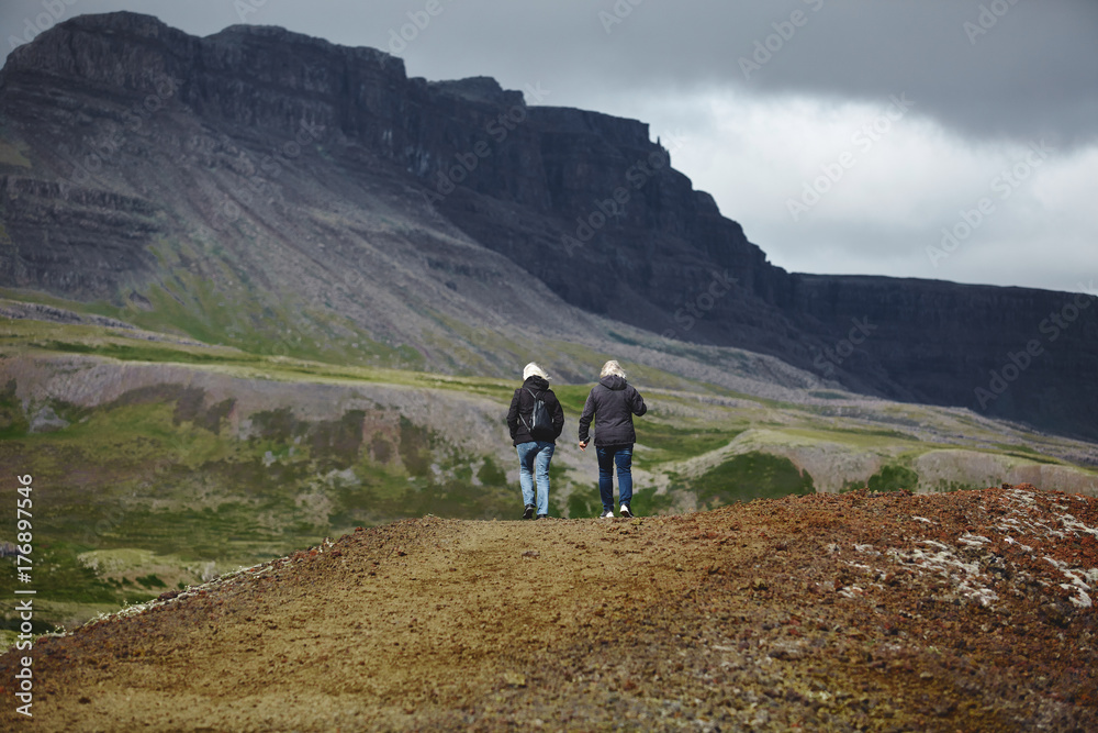 couple adult tourists in waterproof clothes walk along the trail among the lava field in Iceland