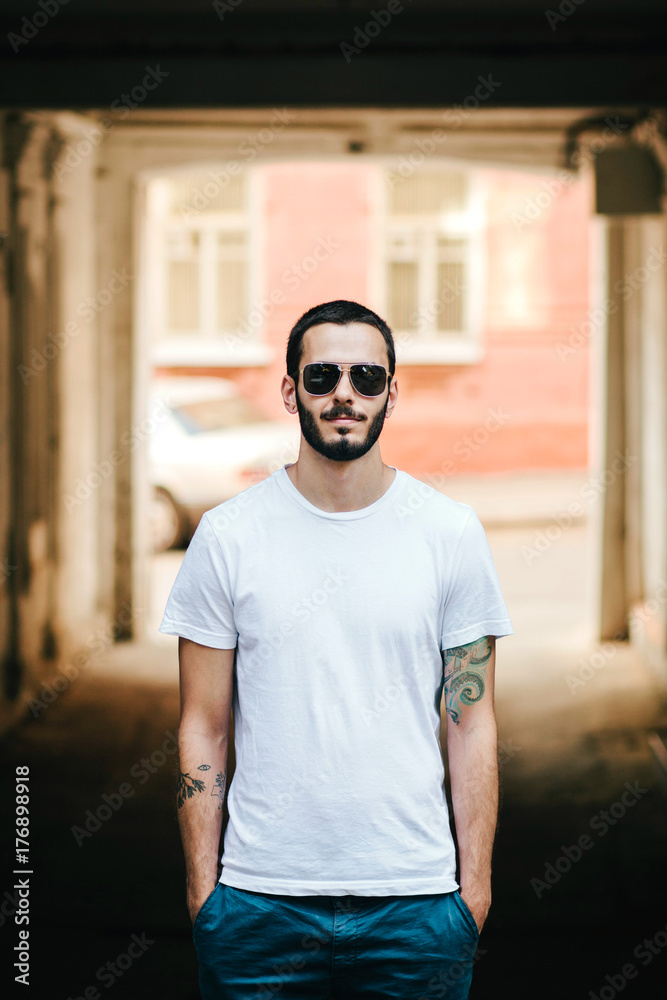 Young stylish man wearing white blank t-shirt with beard in glasses, standing on the street on city background. Street photo Young stylish man wearing white blank t-shirt with beard in glasses, standing on the street on city background. Street photo