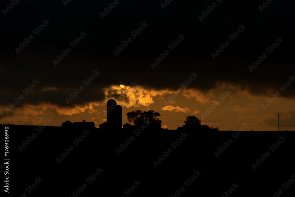 farm buildings and silos are silhouetted by a setting sun