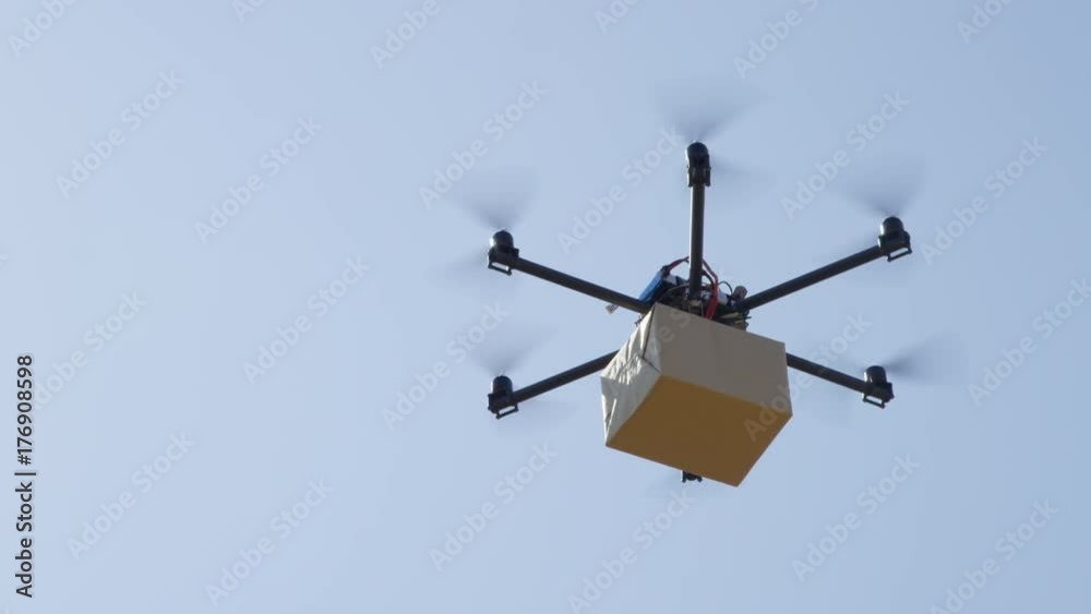 CLOSE UP UAV drone delivery. Multicopter flying big brown package over blue sky. Drone delivering post package to your home. Futuristic shipment by helicopter drone. Multirotor logistics and transport