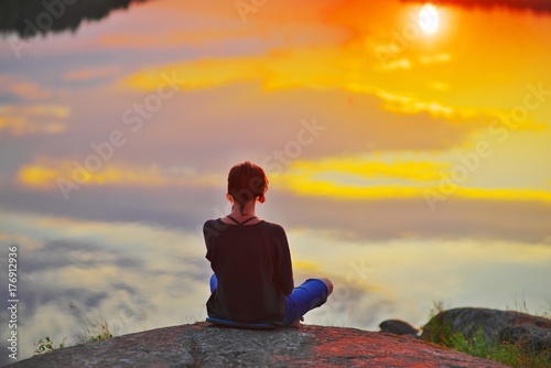 Young woman sitting on the stone enjoying peaceful moment of sunset. In the reflection of the lake water sees clouds and sun. photo