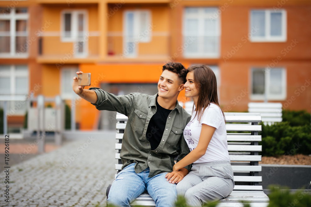 Young couple in shopping. Handsome young couple sitting on the bench and taking a selfie with a mobile phone. Young couple in shopping. Handsome young couple sitting on the bench and taking a selfie with a mobile phone.