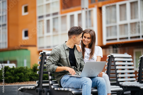 Happy young couple using laptop while sitting on bench in park and smiling each other photo
