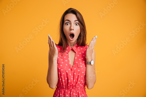 Photo of pretty shocked emotionally woman in red dress looking at camera with opened mouth, isolated on yellow background