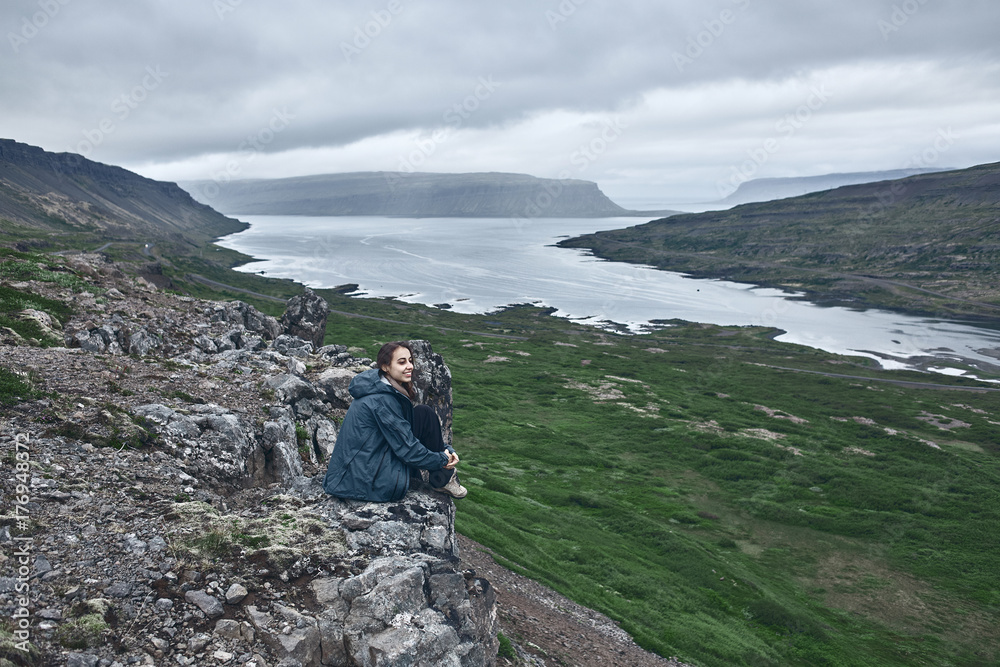 Girl in warm clothing sits on the cliff on background of sea of Iceland.