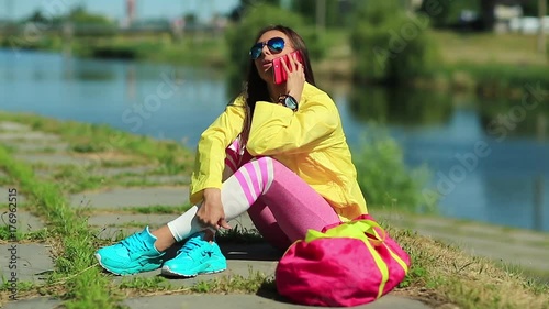 Pretty girl in sunglasses sits near pond and talks via smartphone. Sportswoman in yellow sports jacket sits near lake and speaks on cell phone. Woman in sportswear with mobile phone on the nature photo