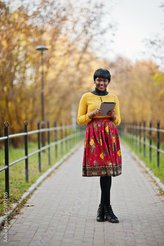 African american girl at yellow and red dress at golden autumn fall park with tablet at hands.