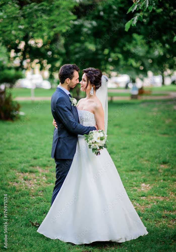 Handsome groom and pretty bride with white bouquet stand on green lawn Handsome groom and pretty bride with white bouquet stand on green lawn