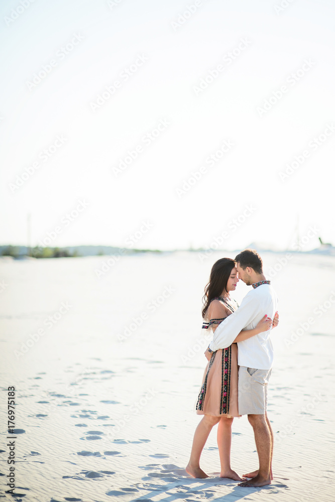 Barefooted couple in bright embroidered clothing hugs tender on a white sand Barefooted couple in bright embroidered clothing hugs tender on a white sand