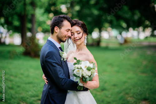 Handsome groom and pretty bride with white bouquet stand on green lawn