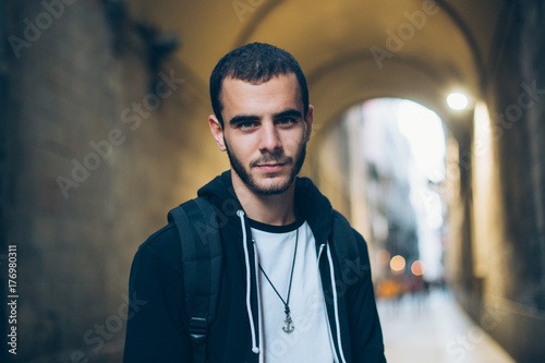 Handsome attractive young man or teenager stands in middle of street in old town, looks straight at camera, serious but slightly smiling in cool and millennial manner photo