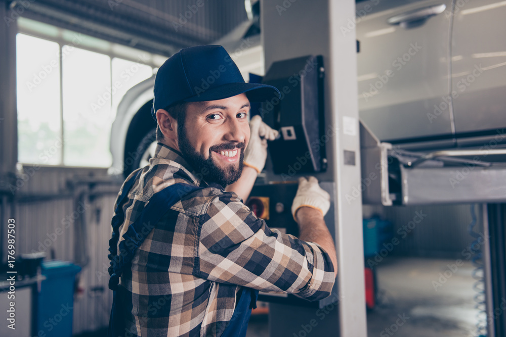 Closeup of cheerful brunet bearded expert in shirt and uniform, hat headwear, pressing the arm lever, car is lifted up, garage interior, work place, station. Engine, motor, transmission check concept Closeup of cheerful brunet bearded expert in shirt and uniform, hat headwear, pressing the arm lever, car is lifted up, garage interior, work place, station. Engine, motor, transmission check concept