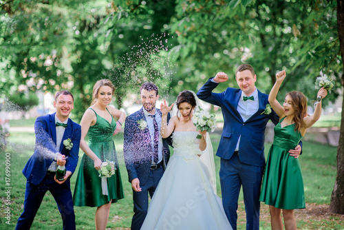 Newlyweds and their friends open champagne bottle standing on a green lawn
