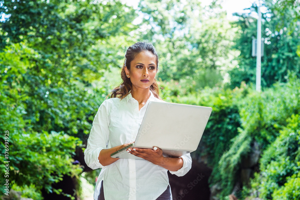 Young Beautiful East Indian American Woman traveling, working in New York, wearing white shirt, holding laptop computer, walking at Central Park. Street bridge with green leaves on background.. Young Beautiful East Indian American Woman traveling, working in New York, wearing white shirt, holding laptop computer, walking at Central Park. Street bridge with green leaves on background..