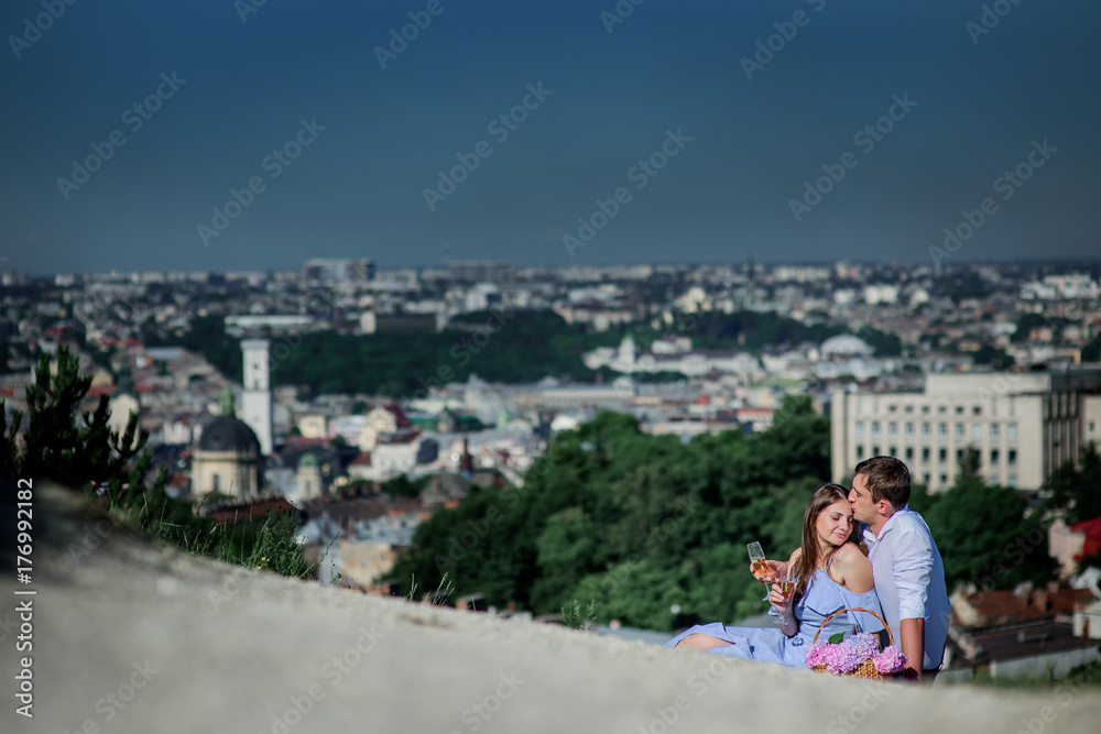 Man and woman drink champagne sitting on the plaid with great cityscape behind them