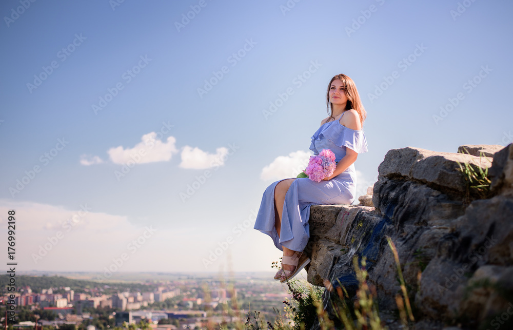 Lady in blue dress sits on the rock with pink bouquet in her arms