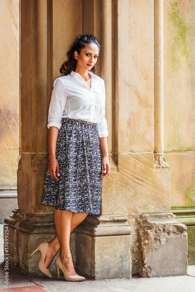 Portrait of Young Beautiful East Indian American Woman in New York, wearing white shirt, black and white patterned skirt, high heals, standing by vintage column, turning around, looking at you.. Portrait of Young Beautiful East Indian American Woman in New York, wearing white shirt, black and white patterned skirt, high heals, standing by vintage column, turning around, looking at you..