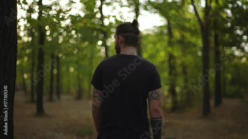 view from back stylish tattooed young man with trendy haircut wearing black T-shirt walking through forest looking around in slow motion