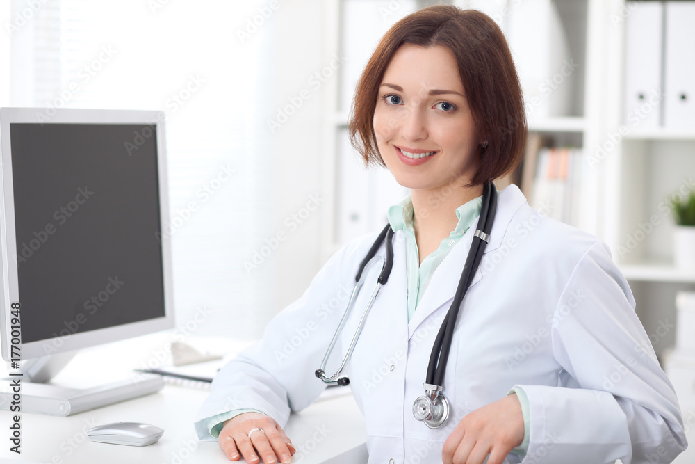 Young brunette female doctor sitting at a desk and working on the computer at the hospital office. Young brunette female doctor sitting at a desk and working on the computer at the hospital office.