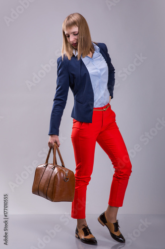 Fashion look of woman dressed red pants, blue jacket, brown bag. model looks at a leather bag. full height photo