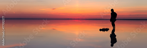 Silhouette of alone man looking toward vibrant sunset reflected in shallow waters of solt lake. Banner size © marmoset