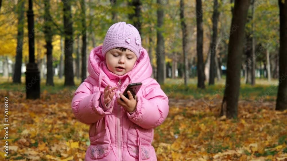 A child in an autumn park with a mobile phone. Little girl is playing with the phone in nature. A child in an autumn park with a mobile phone. Little girl is playing with the phone in nature.