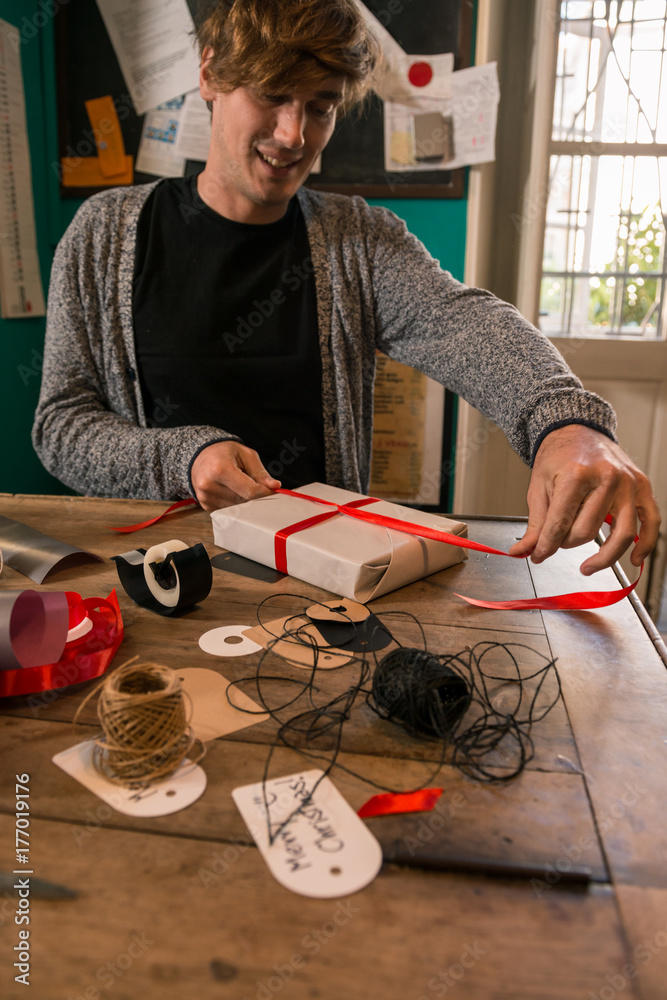 Φωτογραφία Young adult man with long brown hair wrapping christmas present with red ribbon on wooden table in cozy apartment indoor