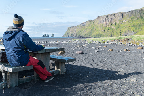 Hombre joven sentado en una mesa de picnic la playa de arena negra de Vik con las formaciones rocosas Reynisdrangur al fondo. Islandia