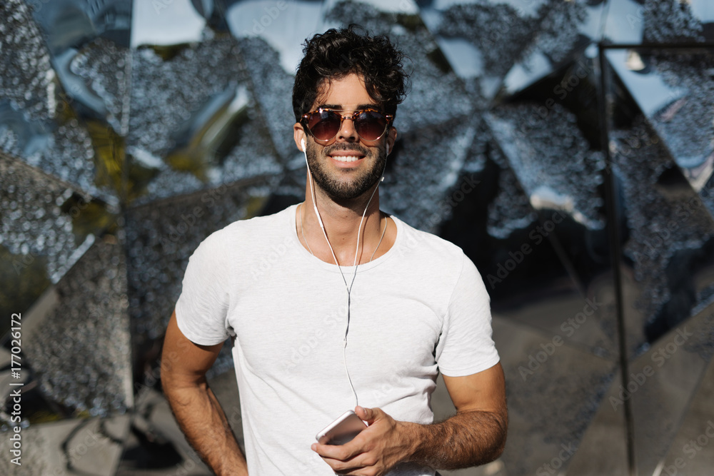 Young man in a blank white t-shirt enjoys the music he listens on a mobile phone while walking outdoors in the morning. Bearded guy wearing sunglasses with a smartphone in his hands. Young man in a blank white t-shirt enjoys the music he listens on a mobile phone while walking outdoors in the morning. Bearded guy wearing sunglasses with a smartphone in his hands.