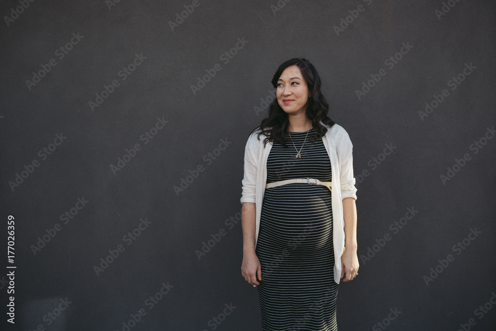Pretty young pregnant woman standing near grey wall outside smiling Pretty young pregnant woman standing near grey wall outside smiling