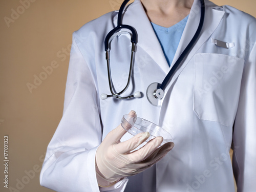 A female doctor holds a petri dish in her hands. A female doctor with a stethoscope on a beige background holds a petri dish.