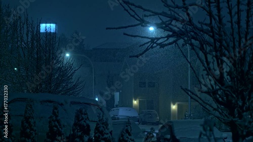 Night time snowfall in a city on peaceful, calm, tranquil evening during winter holidays season on streets of residential suburbs with houses, lights and cars covered with snow by thick snowflakes