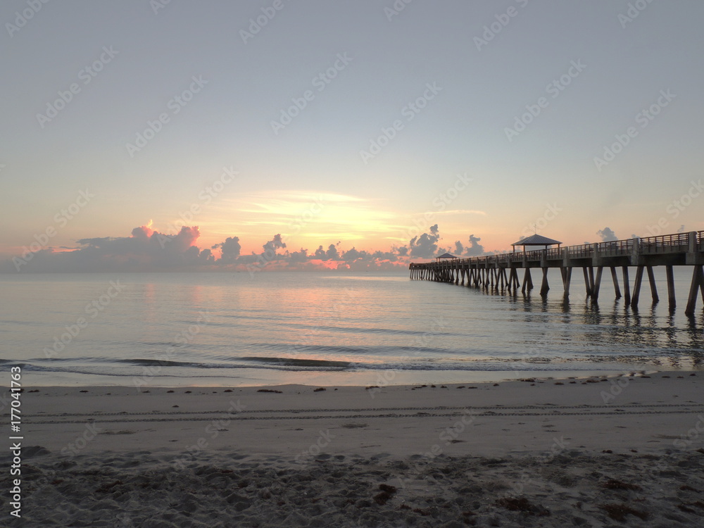 Standing on a sandy beach next to a pier stretching out to a sunrise on the cloudy horizon