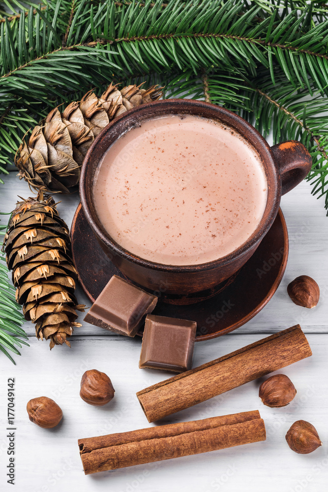 Christmas cup of hot cocoa with milk, broken chocolate cubes, hazelnut and cinnamon sticks, christmas tree branches, pine cone on white wooden planks Christmas cup of hot cocoa with milk, broken chocolate cubes, hazelnut and cinnamon sticks, christmas tree branches, pine cone on white wooden planks