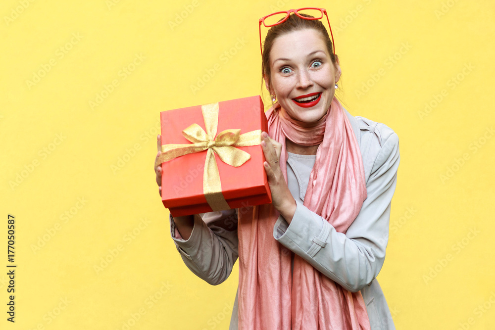 Young adult woman holding gift box and looking at camera and toothy smile.