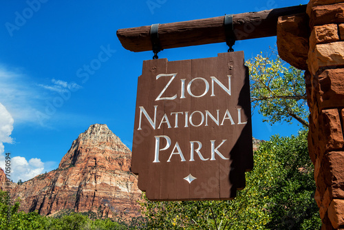 Zion National Park entrance sign: hanging wood panel with blue sky, mountains and trees in background. 