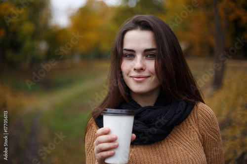 Young woman wearing knitted sweater walking in the autumn park and drinking take away coffee in paper cup. Breakfast on the go, warming concept
