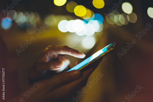 Girl pointing finger on screen smartphone on background illumination glow bokeh light in night atmospheric city, hipster using in hands mobile phone, mockup lights street, online internet concept