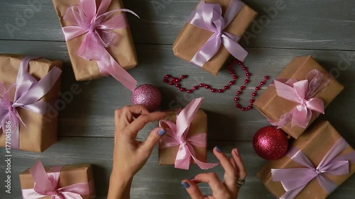 A top-down plan. Fully visible the table with the decorations. Female hands put and finalize Christmas gift wrapped in craftool paper on a wooden table. Bandaging tape and tied in a bow. photo