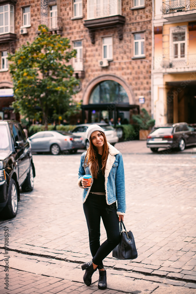 young beautiful brunette drinking coffee walking around the city.denim jacket,urban backpack Posing against the window of the boutique Model looking and drinking coffee