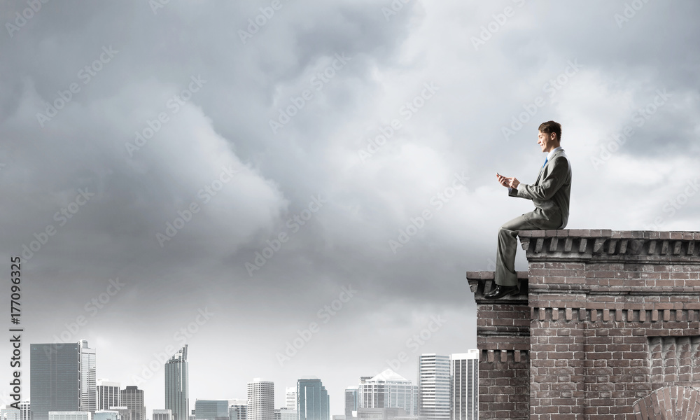 Man on roof edge send message with smartphone and cityscape at background Man on roof edge send message with smartphone and cityscape at background