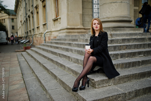 Elegant blonde girl wear on black posing at streets of town, sitting on stairs with cup of coffee at hands. © AS Photo Family