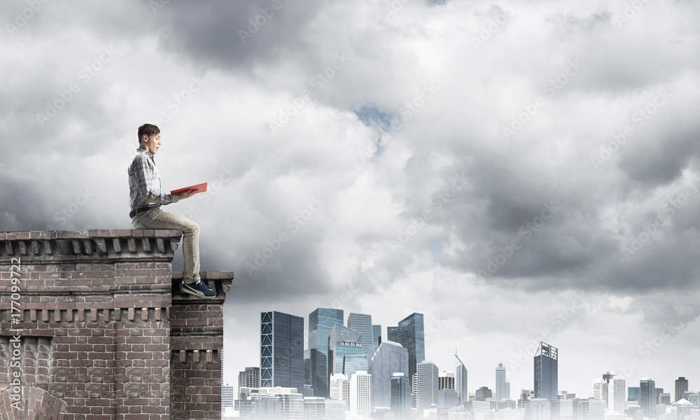 Man on roof edge reading book and cityscape at background