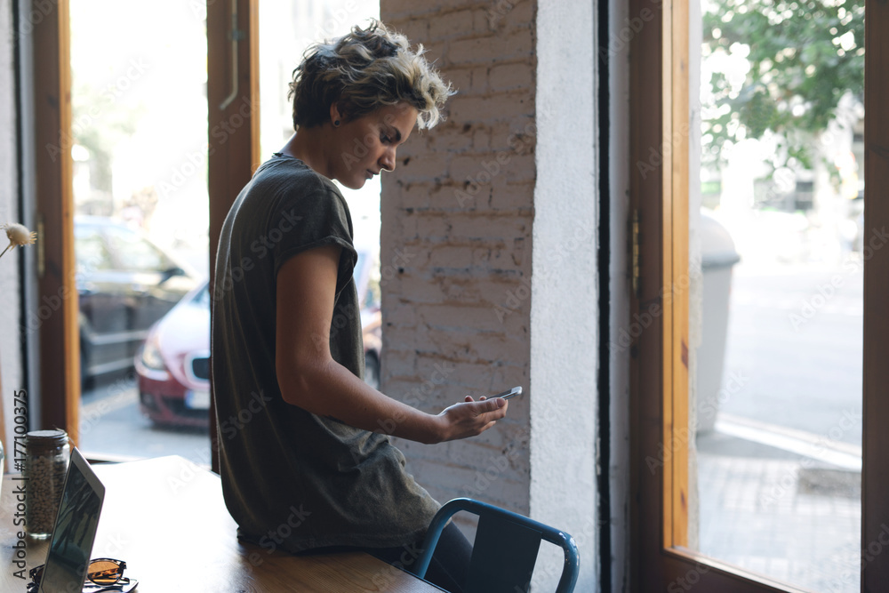 Beautiful blonde woman wearing casual clothes is reading emails on a smartphone while standing indoors beside a bid window. Hipster girl is chatting with friends by social networks on a mobile phone.