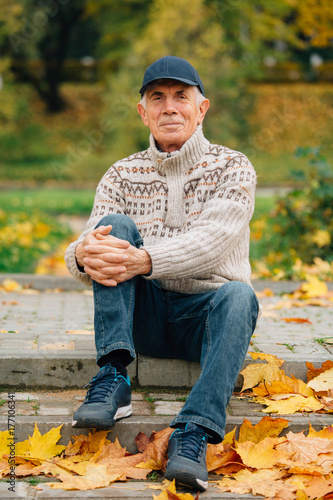 Portrait of pretty senior man in cap sitting in colorful autumn park. Pensioner resting outdoors. Beutiful fall.