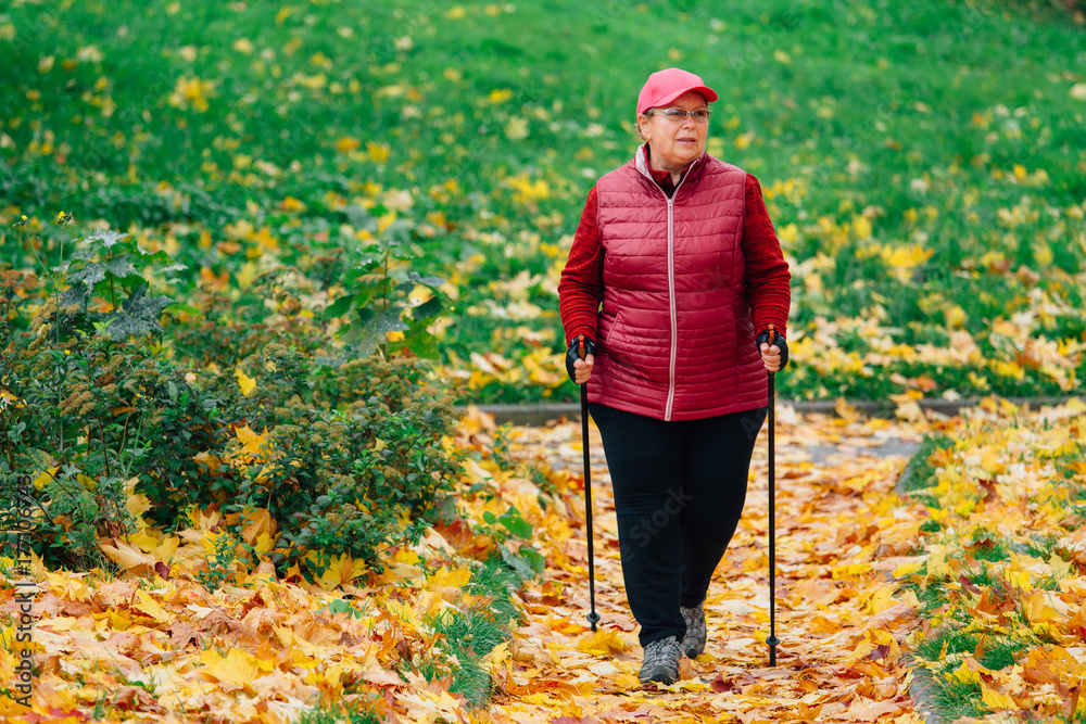Senior woman with nordic walking poles in colorful autumn park. Healthy life concept. Old sportswoman doing exercise outdoors.