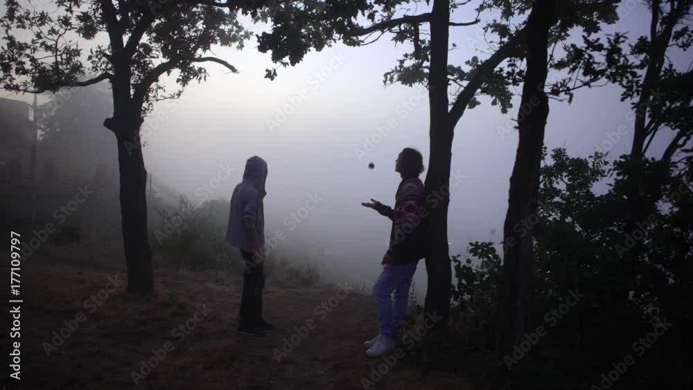 from above young slim man throwing up hacky sack standing under trees on hill over sea covered with fog with male friend looking into distance slowmotion from above young slim man throwing up hacky sack standing under trees on hill over sea covered with fog with male friend looking into distance slowmotion